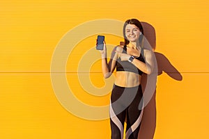 Young satisfied beautiful sporty woman in black sportwear standing near orange wall background and holding phone, pointing finger