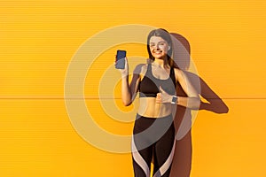 Young satisfied beautiful sporty woman in black sportwear standing near orange wall background and holding phone, showing thumbs