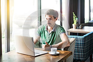 Young satisfied businessman in green t-shirt sitting and working on laptop near window and showing thumbs up like sign