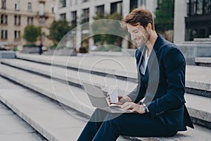 Young satisfied businessman in stylish suit sitting on stairs outside with laptop and coffee to go