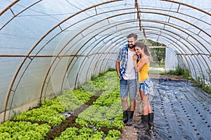 Young satisfied family couple standing in the greenhouse and smiling looking at the vegetables they planted as small business and