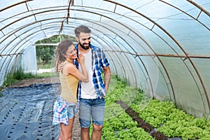 Young satisfied family couple standing in the greenhouse and smiling looking at the vegetables they planted as small business and