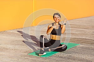 Young satisfied sporty girl in black sporwear in morning on street, sitting on green mat, pointing finger and showing good result