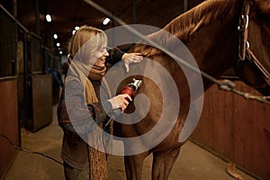 Young satisfied woman grooming horse with electric shaver in ranch stable