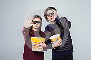 Young scared couple, woman and man in 3d glasses watching movie film on date holding buckets of popcorn covering faces with palm