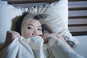 Young scared and stressed Asian Chinese woman lying in bed suffering nightmare in fear and panic grasping blanket covering her hor