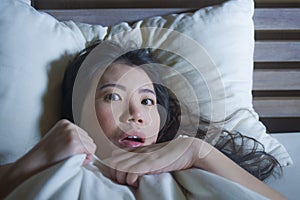 Young scared and stressed Asian Chinese woman lying in bed suffering nightmare in fear and panic grasping blanket covering her hor