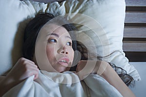 Young scared and stressed Asian Chinese woman lying in bed suffering nightmare in fear and panic grasping blanket covering her hor
