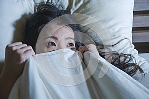 Young scared and stressed Asian Chinese woman lying in bed suffering nightmare in fear and panic grasping blanket covering her hor