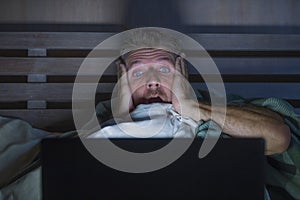 Young scared and stressed man in bed watching internet horror movie late night with laptop computer or bedroom television in