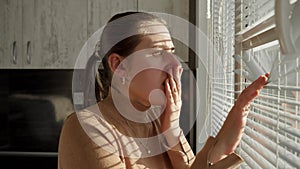 Young scared woman got shocked and terrified after looking outdoors through window blinds.