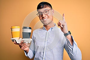 Young scholarship holder business man holding take away coffee over isolated yellow background surprised with an idea or question