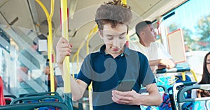 Young school-age boy rides public transportation bus to elementary school holding phone in hand, looks
