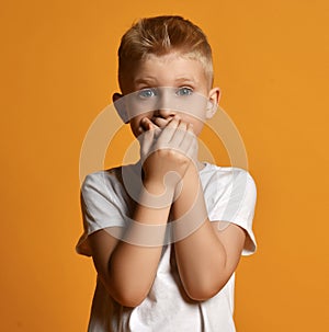 Young school boy kid in white t-shirt stands scared, eyes wide opened in surprise, covering his mouth with both hands