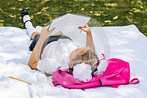 A young schoolgirl does homework while lying on a backpack in a sunny autumn park. Outdoor education for children. Back to school