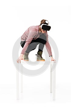 Young screaming man standing on white table in studio while playing with virtual reality glasses on white background
