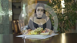 young seductive brunet woman in black dress with cleavage eating fruits grapes alone at table in fancy restaurant