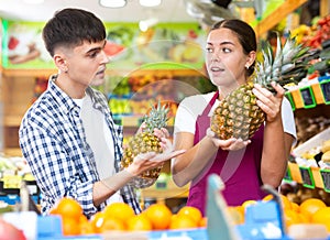 Young seller girl in apron selling pineapple to man customer in grocery store