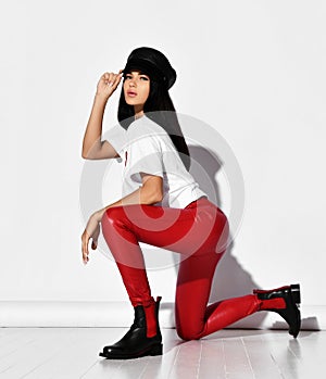 Young sensual woman vamp in tight red leather pants, white t-shirt and leather cap standing on her knee side to camera