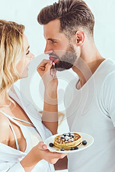 Sensual woman feeding boyfriend with strawberry while holding plate with pancakes
