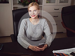 Young serious adorable blonde business lady posing in home office room sitting at the desk with notebook holding mobile phone