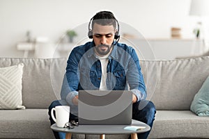 Young serious Arab man wearing headphones, using laptop computer, working online from home