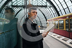 Young serious Asian businessman with newspaper confidently looking in camera while waiting train at subway station