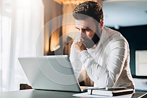 Young serious bearded businessman standing in office near table and using laptop. Man works on computer, checks e-mail