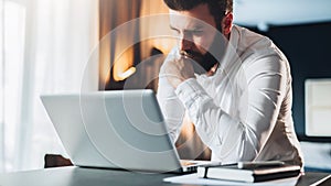 Young serious bearded businessman standing in office near table and using laptop. Man works on computer, checks e-mail