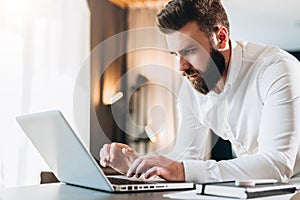 Young serious bearded businessman standing in office near table and using laptop. Man works on computer, checks e-mail