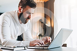 Young serious bearded businessman standing in office near table and using laptop. Man works on computer, checks e-mail