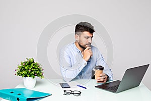 Young serious bearded businessman working on computer at table,drinking coffee. Man analyzes information, data, develops business