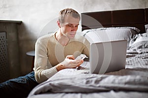 A young serious businessman is sitting at home near the bed. Freelancer works at home in a comfortable environment. A man is