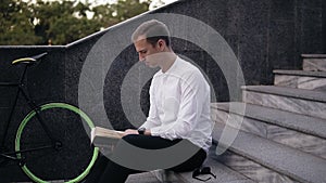 Young serious caucasian man reading book, sitting on city urban stairs, studying or reading, leisure time in city park