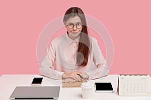 Young serious Caucasian woman wears spectacles, formal shirt, has everything on its place at table, surrounded with