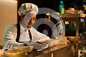 Young serious chef in eyeglasses and uniform reading recipe of new dish