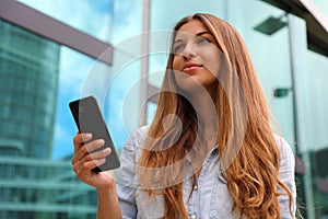 Young serious and determined business woman standing in front of modern building with phone in her hand