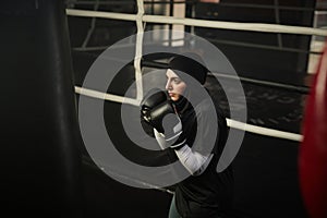 Young serious female boxer in activewear and hijab looking at punching bag