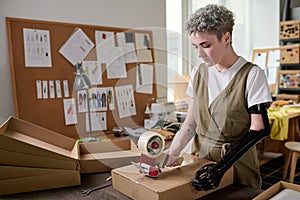 Young serious female worker of warehouse sealing packed box with cellotape