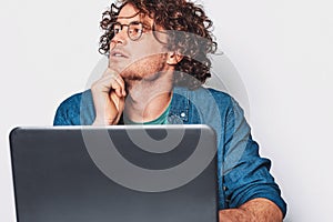 Young serious man with curly hair sitting at table working at laptop. Businessman wearing round glasses and working on laptop