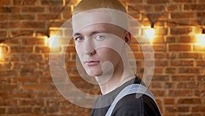 Young serious man hipster is turning his head, watching at camera, brick background, side view