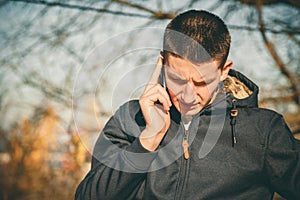 Young serious man holding mobile phone, using smartphone, making a call, talking on the phone, standing on sunny street