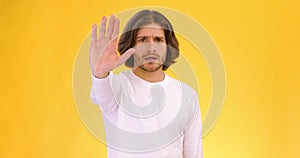 Young serious man showing stop gesture and crossing hands, shaking head in rejection, orange studio background