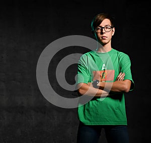 Young serious teen boy in green t-shirt, jeans and glasses standing and looking camera over dark background