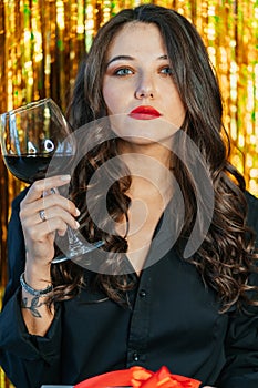 Young serious woman with make-up wearing black shirt, raising glass with red wine on golden tinsel background. New year.