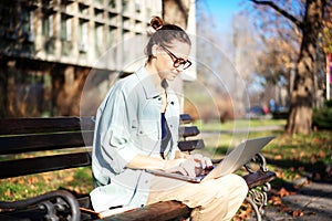 A young serious woman student sitting on a bench in the park and typing on her laptop on a sunny day in universiry campus