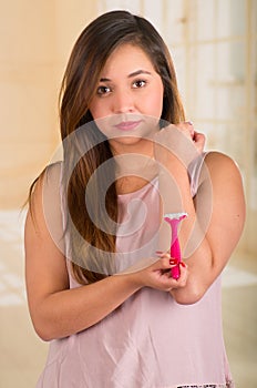 Young serious woman using a shaver in her arm, with a bath background