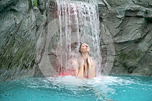 Young sexy blond woman in pink bikini enjoys the falling water of the waterfall