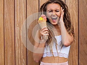Young blonde girl with dreads eating multicolored ice cream in waffle cones in summer evening, cries holding her head. photo