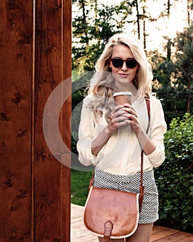 Young blonde girl with long hair in sunglasses with brown vintage bag holding a cup of coffee have fun and good mood looking